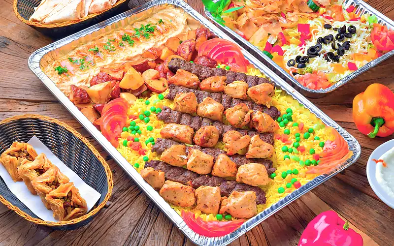 Family tray of beef kabab and chicken shish tawook over yellow rice with garlic potatoes and hummus, with a salad tray behind