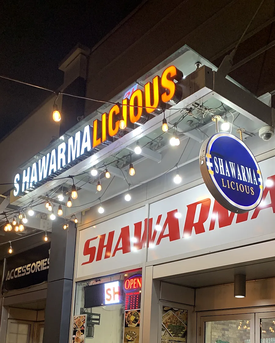 Shawarmalicious sign and round badge lit up at night on 17th Ave SW