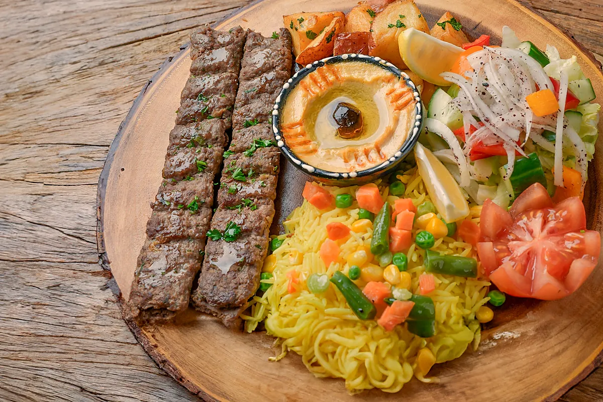 Two beef kabab skewers on a wooden board with rice, hummus and salad