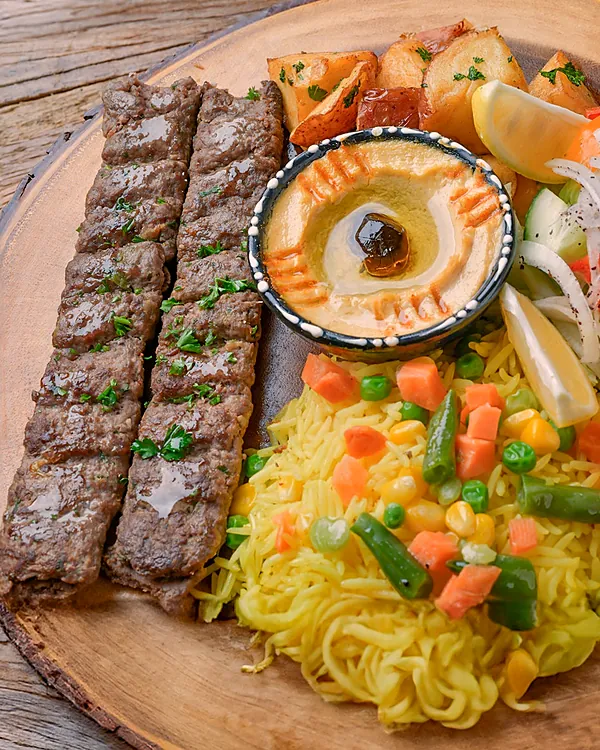 Two beef kabab skewers on a wooden board with rice, hummus and salad